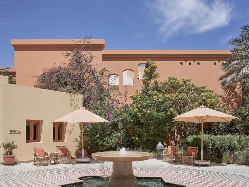 Innenhof mit Springbrunnen, Sonnenschirmen, Pflanzen und einem rosafarbenen Gebäude unter blauem Himmel.