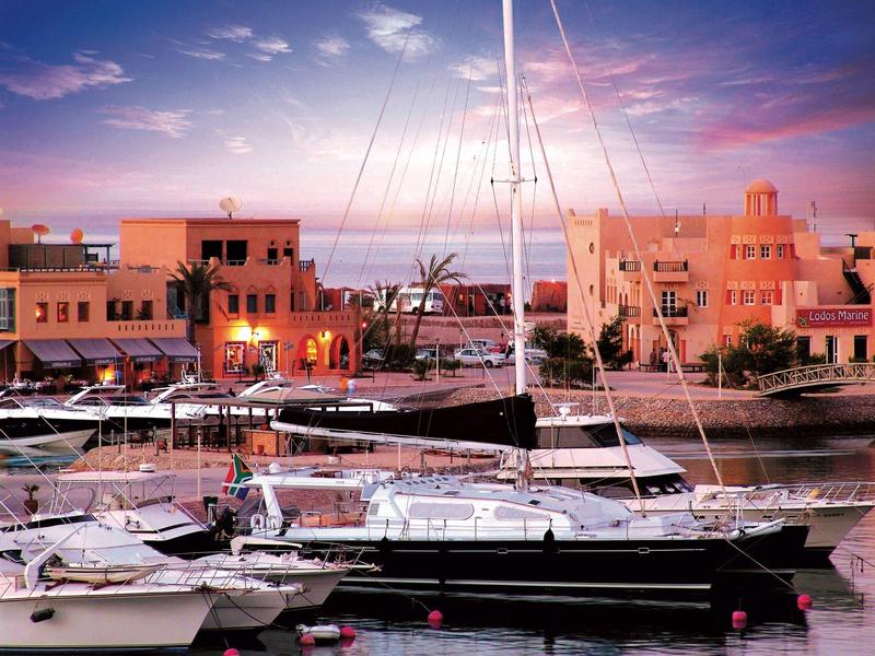 Yachten liegen ruhig im Hafen vor orangefarbenen Gebäuden bei Sonnenuntergang.