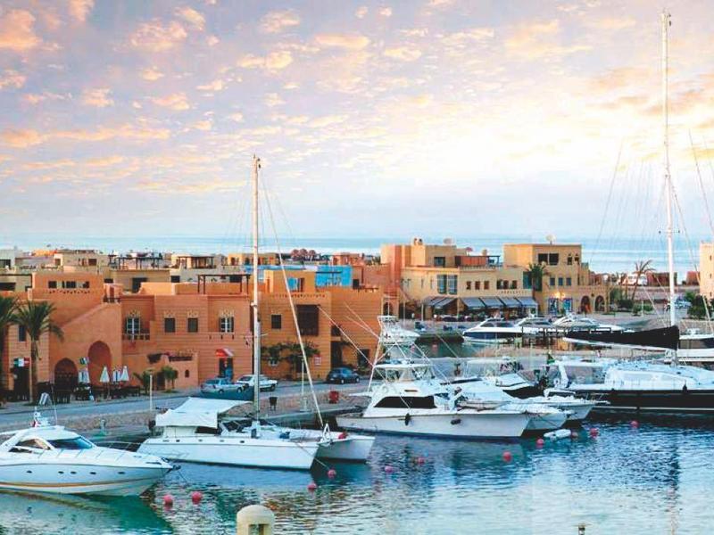 Hafen mit mehreren Booten und Segelyachten, im Hintergrund niedrige Häuser und bewölkter Himmel.