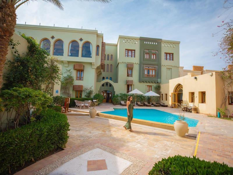 Innenhof mit Schwimmbad, Palmen, Pflanzen und einem mehrstöckigen Gebäude im Hintergrund.