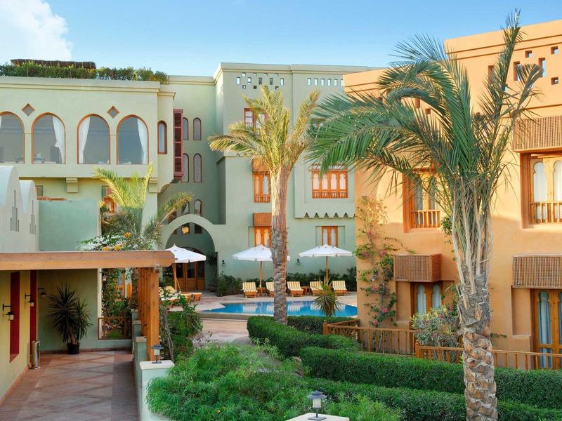 Bunter Gebäudekomplex mit Palmen, grünen Sträuchern und blauem Pool unter hellem Himmel.