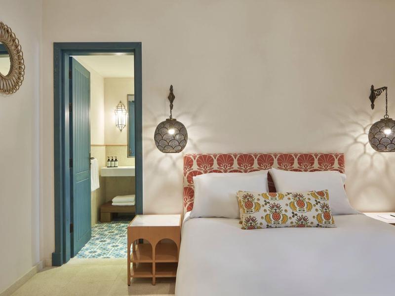 Schlafzimmer mit weißem Bett, gemustertem Kopfteil, Nachttischen und blauem Türrahmen zum Bad.