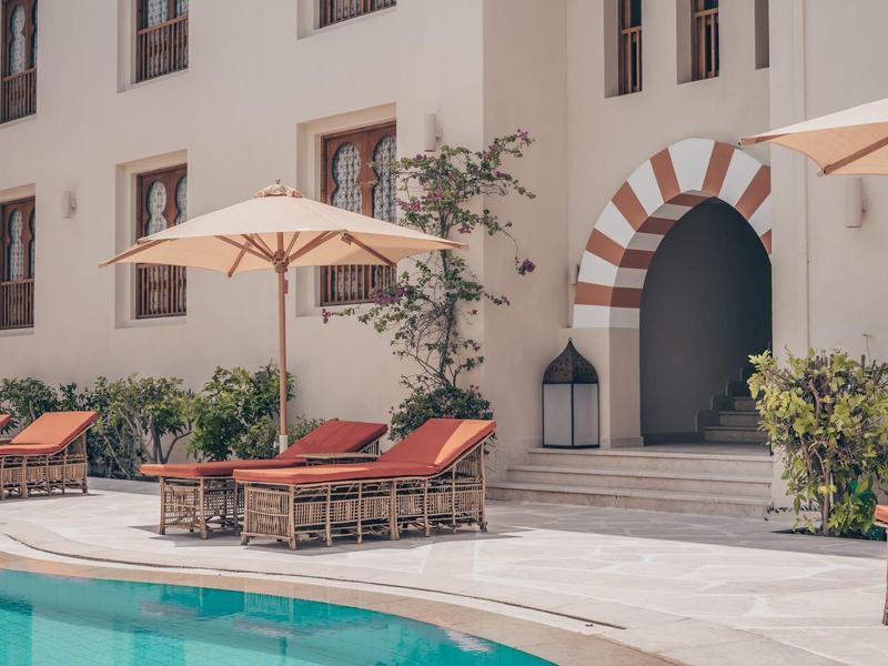Pool mit Orangetönen Sonnenliegen, Sonnenschirmen und Eingang mit Rundbogen in weißem Gebäude.