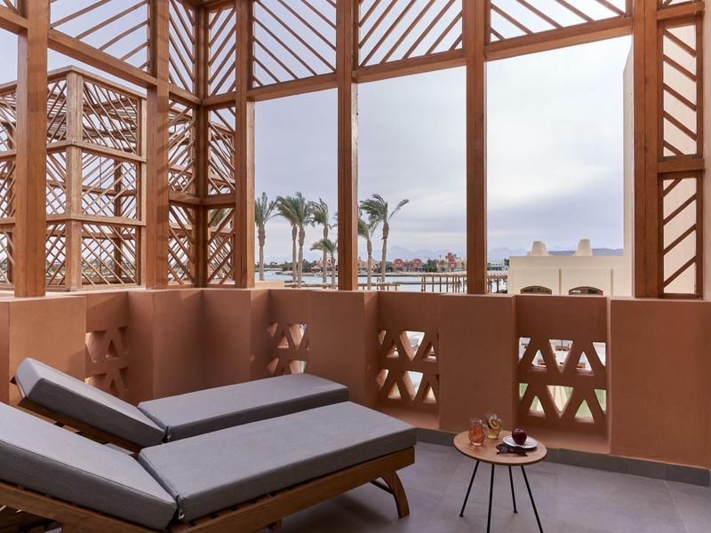 Gemütlicher Balkon mit zwei grauen Liegestühlen, braunen Holzelementen und Palmenblick am Meer.