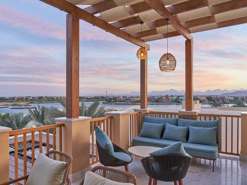 Überdachte Terrasse mit blauen Sitzmöbeln, Holzgeländer und Blick auf Palmen und Berge bei Sonnenuntergang.