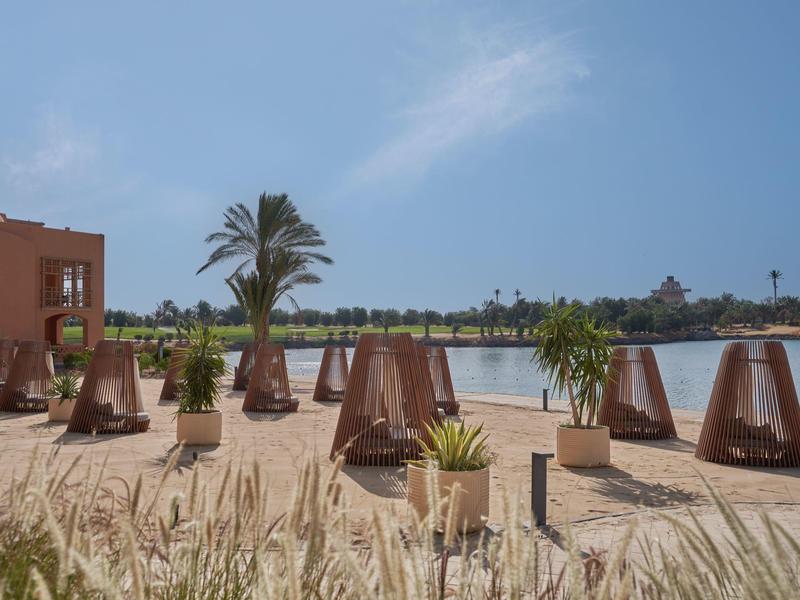 Sandstrand mit mehreren Holzkörben und Palmen vor einem ruhigen See unter blauem Himmel.