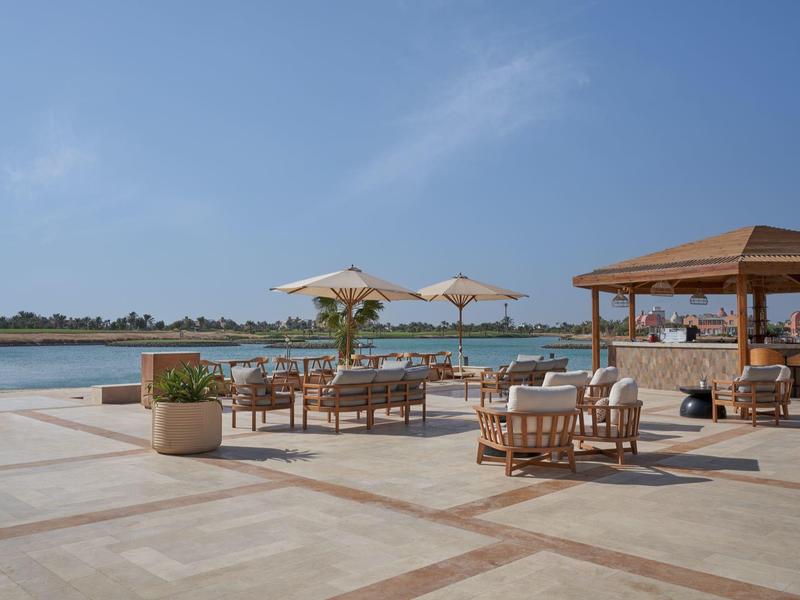 Offene Strandbar mit Holzmöbeln, Sonnenschirmen, Pool und Blick auf ruhiges Meer bei klarem Himmel.