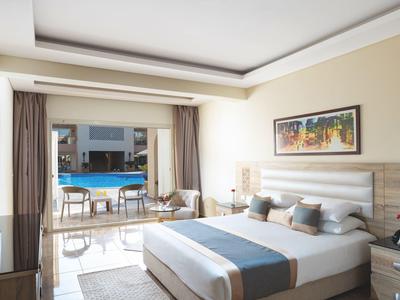 Helles Hotelzimmer mit großem Bett, modernen Möbeln und Blick auf den Außenpool durch eine offene Glasfront.