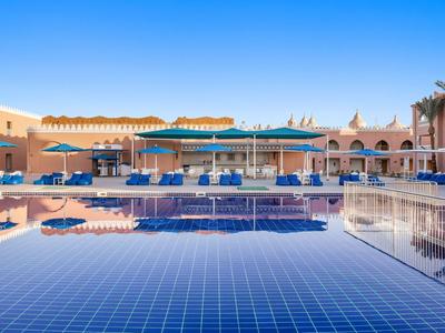 Hotelpool mit blauer Kachel, Liegestühlen und Sonnenschirmen vor einem sonnigen Hotelgebäude.