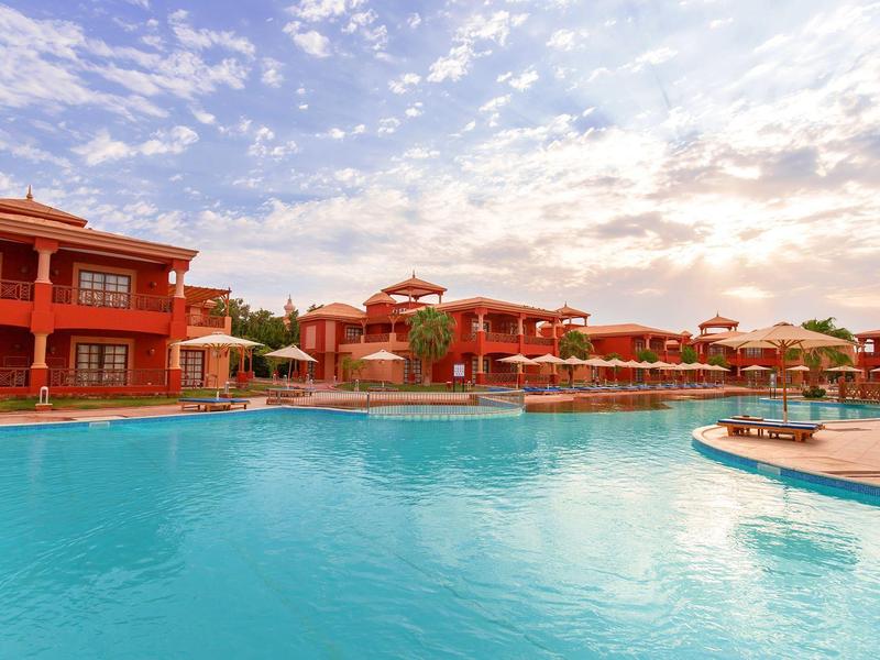 Großer blauer Pool vor roten Gebäuden unter teilweise bewölktem Himmel.