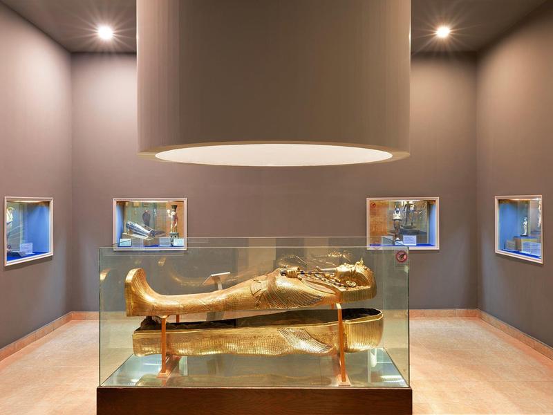 Moderne Galerie mit goldener ägyptischer Sarkophag in Glasvitrine, umgeben von vier kleinen Fenstern.