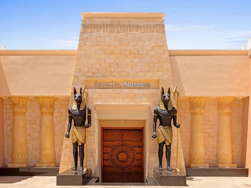 Eingang mit zwei großen Anubis-Statuen und Säulen im ägyptischen Stil vor blauer Himmel.