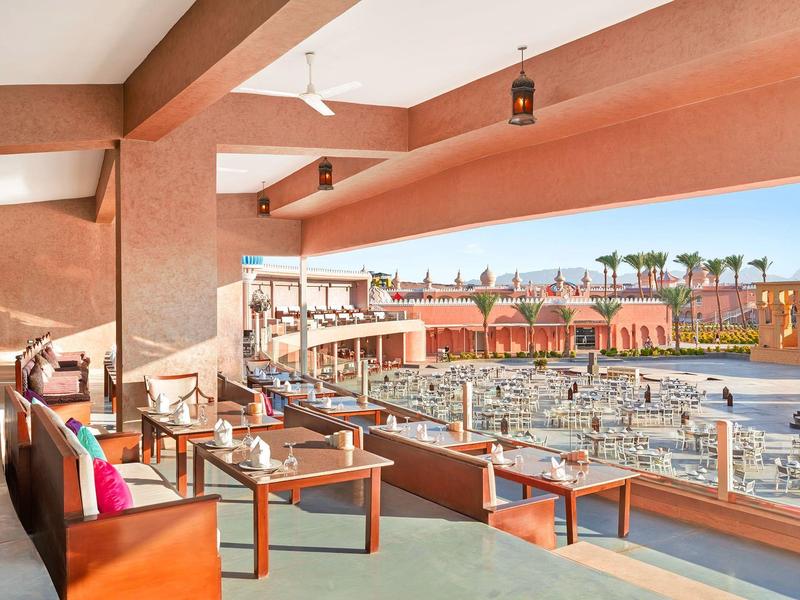 Terrassenbereich mit Bänken, Tischen und Blick auf Pool und Gebäude in warmen Farbtönen.