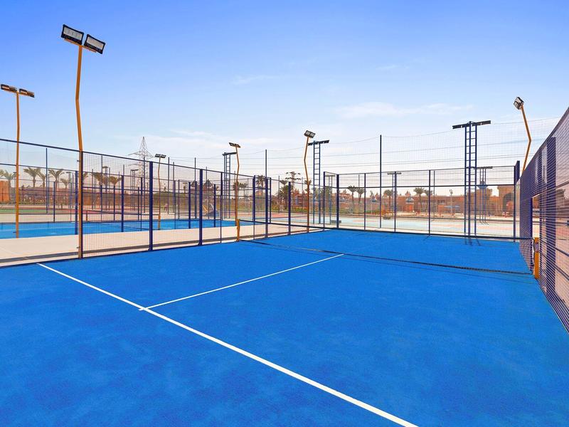 Blauer Paddle-Tennisplatz mit hohen Zäunen und Flutlichtmasten unter klarem Himmel.