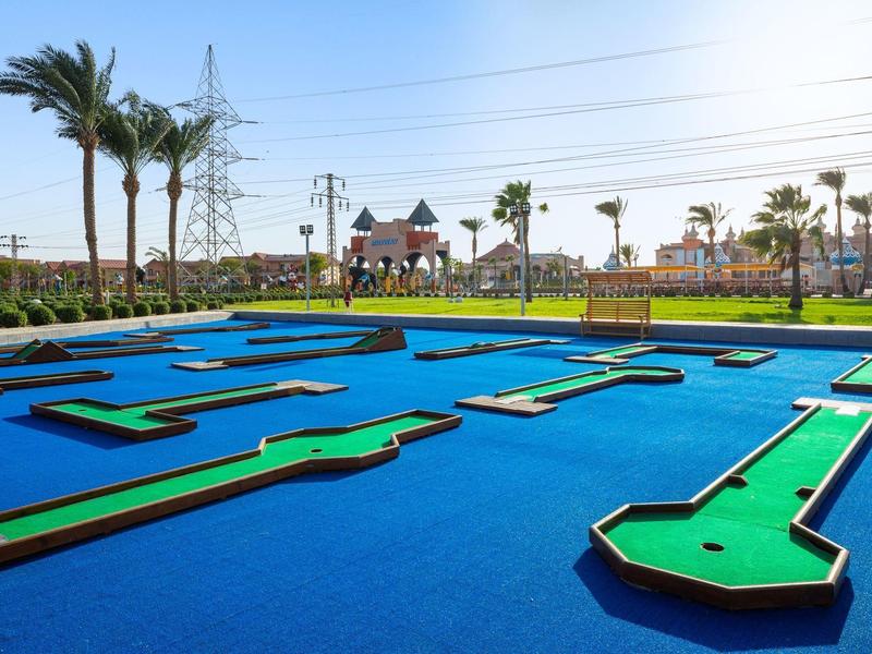 Minigolfplatz mit grünem Kunstrasen und blauer Umrandung, Palmen und Gebäude im Hintergrund.