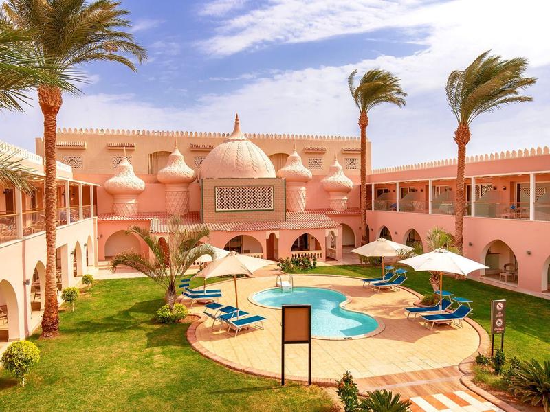 Innenhof eines Hotels mit Pool, Palmen, grüner Wiese und rosa Gebäuden unter blauem Himmel.
