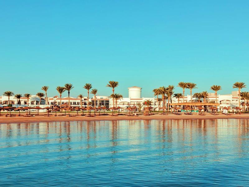 Hotel am Strand mit Palmen und klarem blauem Wasser im Vordergrund.