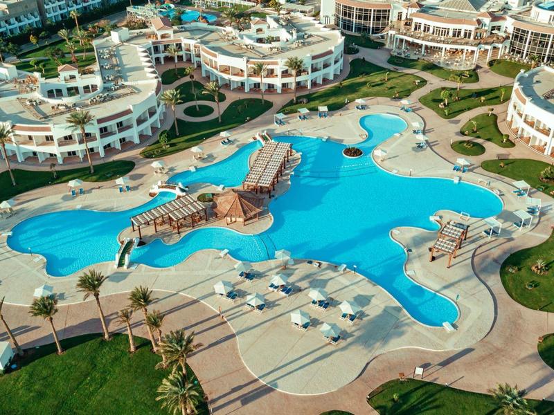 Großer blauer Pool mit Liegestühlen, Palmen und runden Wohngebäuden im Hintergrund.