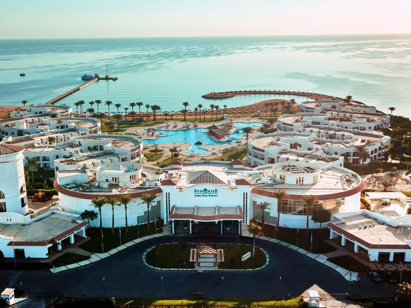 Große Hotelanlage am Meer mit weißen Gebäuden, Pools, Palmen und einem Hafen im Hintergrund.