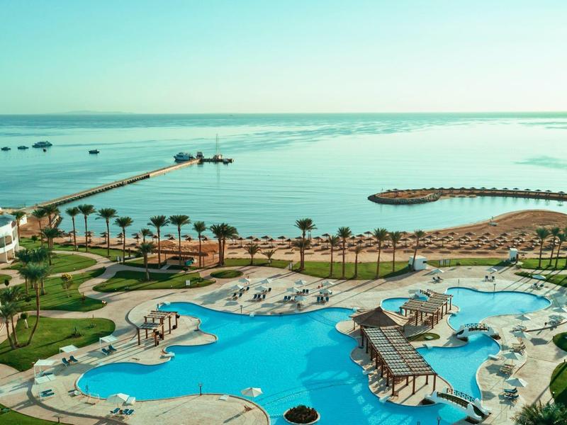 Blick auf großes Hotelpool mit Palmen, Liegen und blauem Meer im Hintergrund bei sonnigem Himmel.