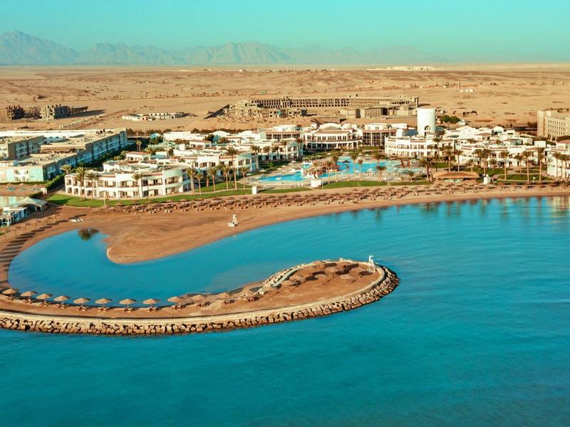 Küste mit halbkreisförmigem Hafen, weißen Gebäuden und bergiger Wüstenlandschaft im Hintergrund.