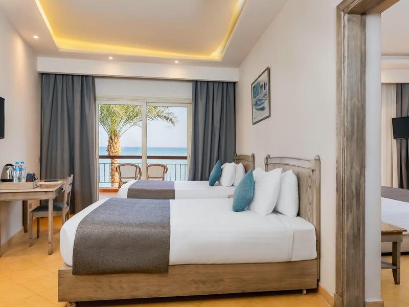 Helles Hotelzimmer mit großem Bett, Holzmöbeln und Meerblick durch große Fenster.