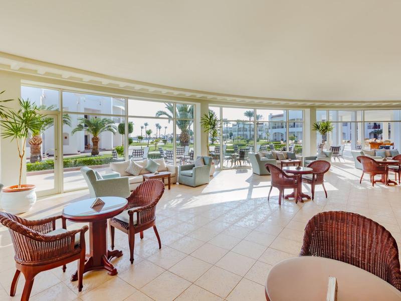 Helle, moderne Lounge mit großen Fenstern, weißen Fliesen und roten Korbstühlen an Rundtischen.