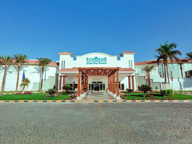 Großes weißes Gebäude mit Palmen und roter Pergola vor blauem Himmel und grauem Pflaster.