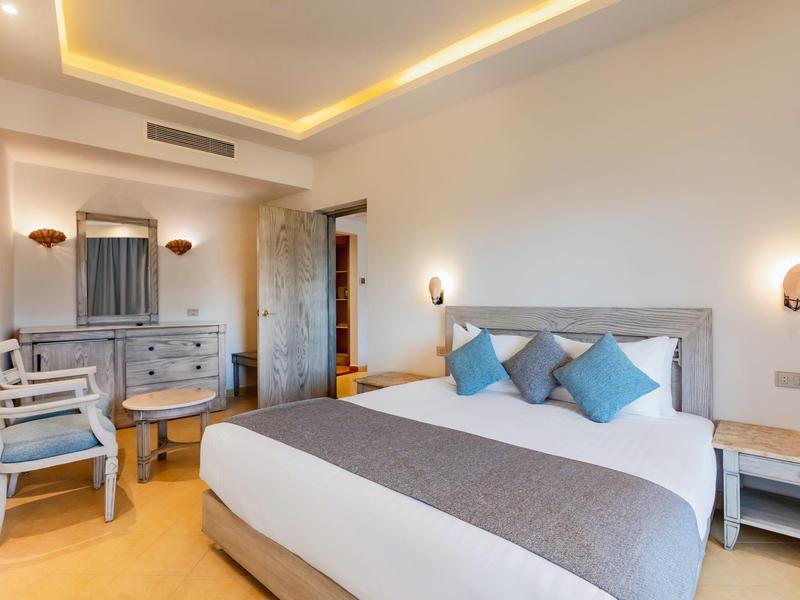 Helles Schlafzimmer mit großem Bett, grauen und blauen Kissen, Holzmöbeln und indirekter Deckenbeleuchtung.