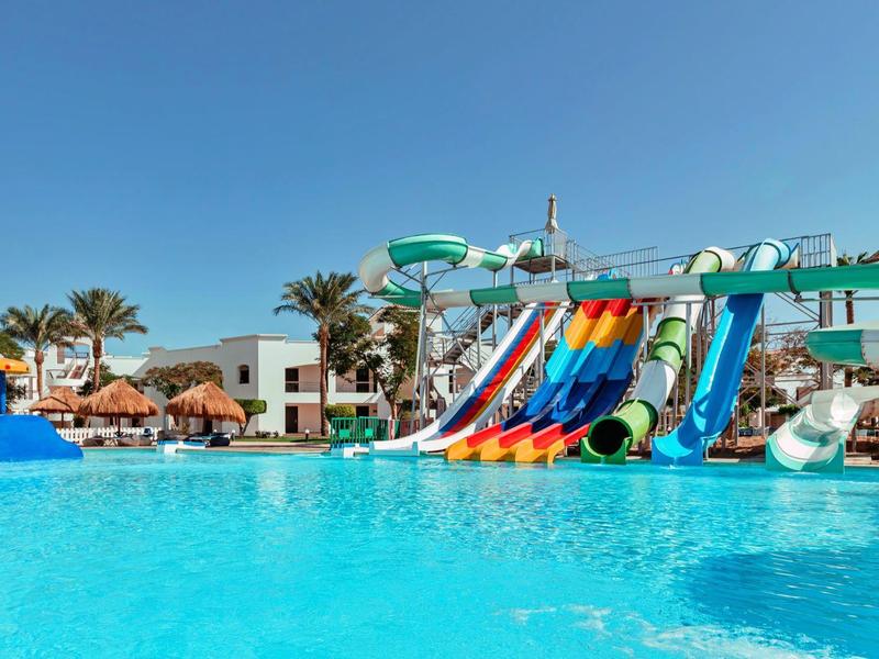 Großer blauer Pool mit bunten Wasserrutschen, Palmen und Sonnenschirmen bei klarem Himmel.