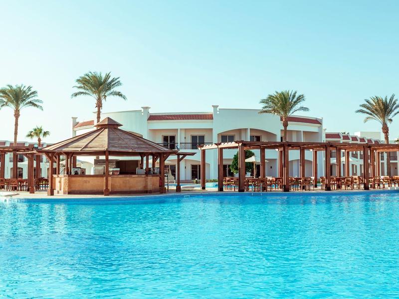 Großer blauer Swimmingpool vor einer weißen Hotelanlage mit Palmen und Holzkonstruktionen.