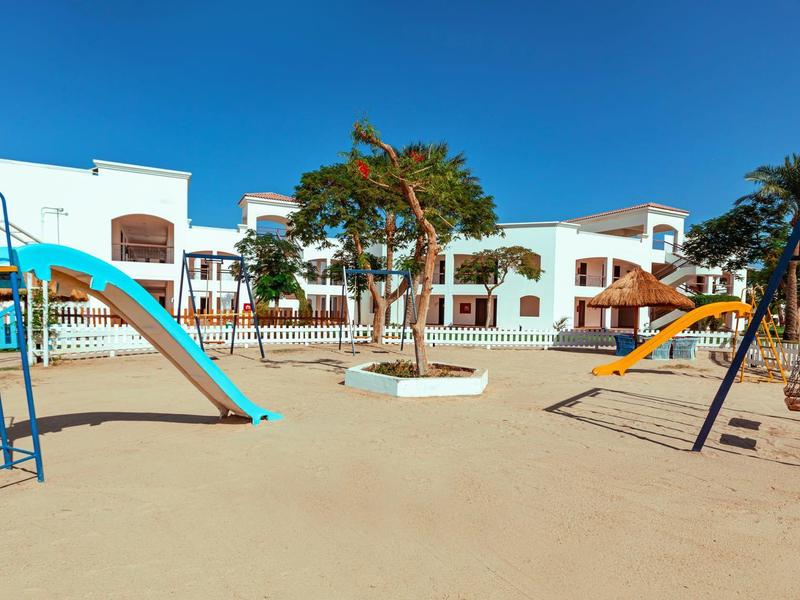 Spielplatz mit Rutschen, Schaukeln und Sandfläche vor weißen Gebäuden und blauem Himmel.
