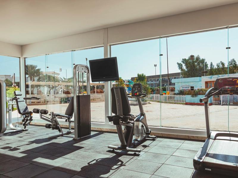 Helles Fitnessstudio mit großen Fenstern, Laufband, Kraftmaschinen und TV-Bildschirm.