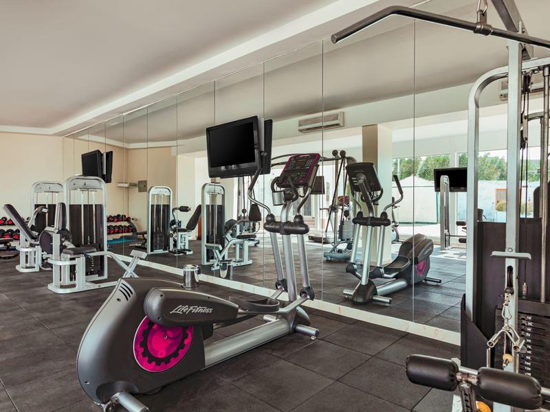 Moderner Fitnessraum mit verschiedenen Trainingsgeräten, Spiegelwand und großen Fenstern.