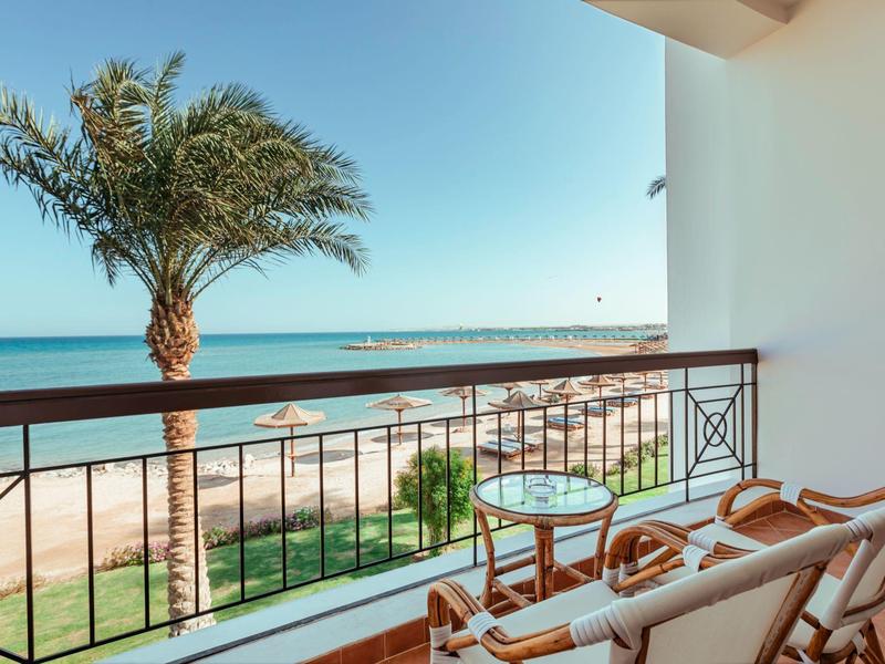 Balkon mit Stühlen und Tisch, Blick auf Palmen, Strand und ruhiges blaues Meer unter klarem Himmel.