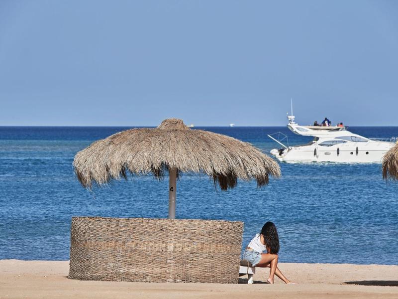 Strand mit Strohmantel Sonnenschirm, Sand, blauem Meer und vorbeifahrendem weißen Boot.