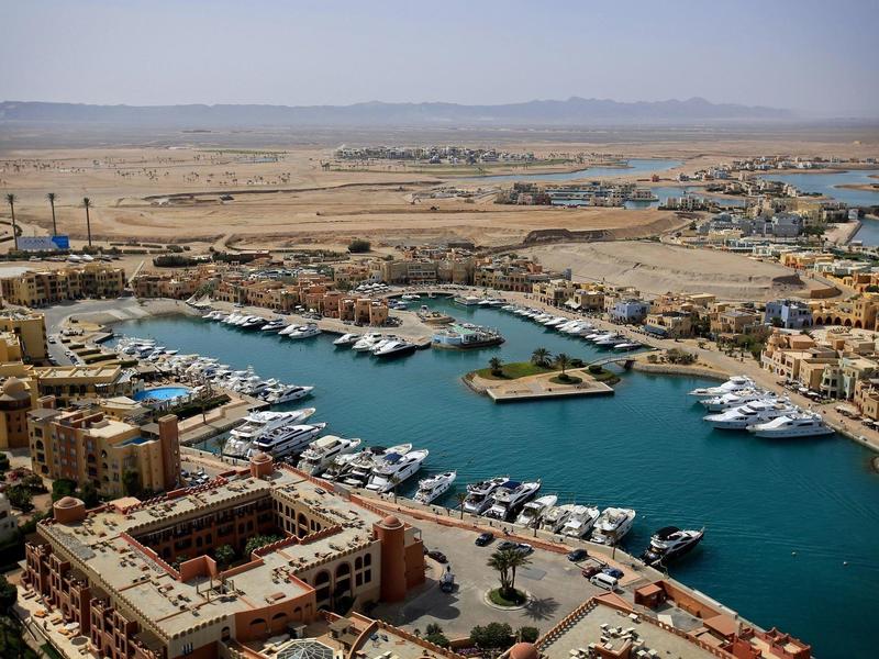 Blick auf einen Hafen mit Yachten, umgeben von Gebäuden und weiter Landschaft mit Bergen am Horizont.