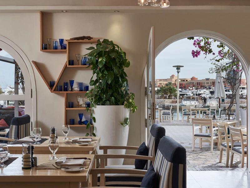 Innenraum eines modernen Restaurants mit Holzmöbeln, großen Fenstern und Blick auf Terrasse und Hafen.