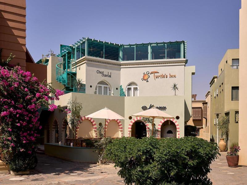 Hellbeige Gebäude mit terrakottafarbenen Akzenten, blühenden Sträuchern und blauer Himmel im Hintergrund.