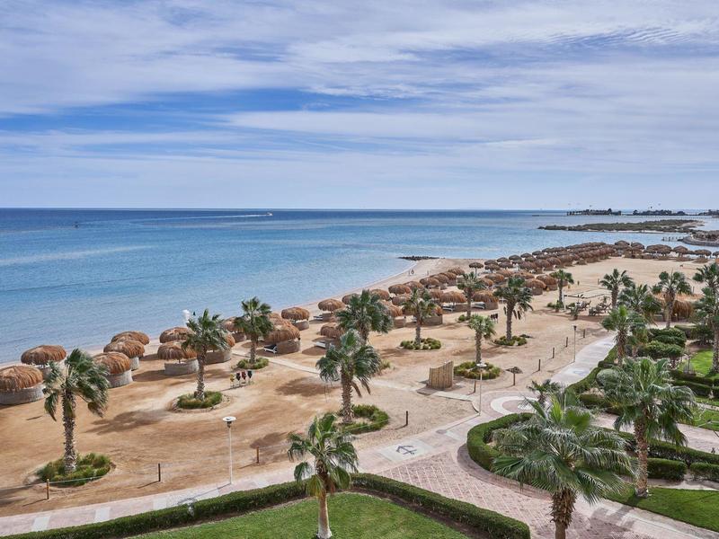 Strand mit Palmen, Sonnenschirmen aus Stroh und blauem Meer unter leicht bewölktem Himmel.