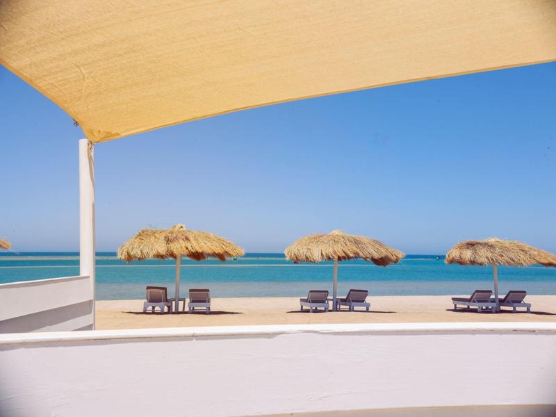 Strand mit drei Strohhut-Sonnenschirmen, Liegen und klarem blauem Meer unter blauem Himmel.