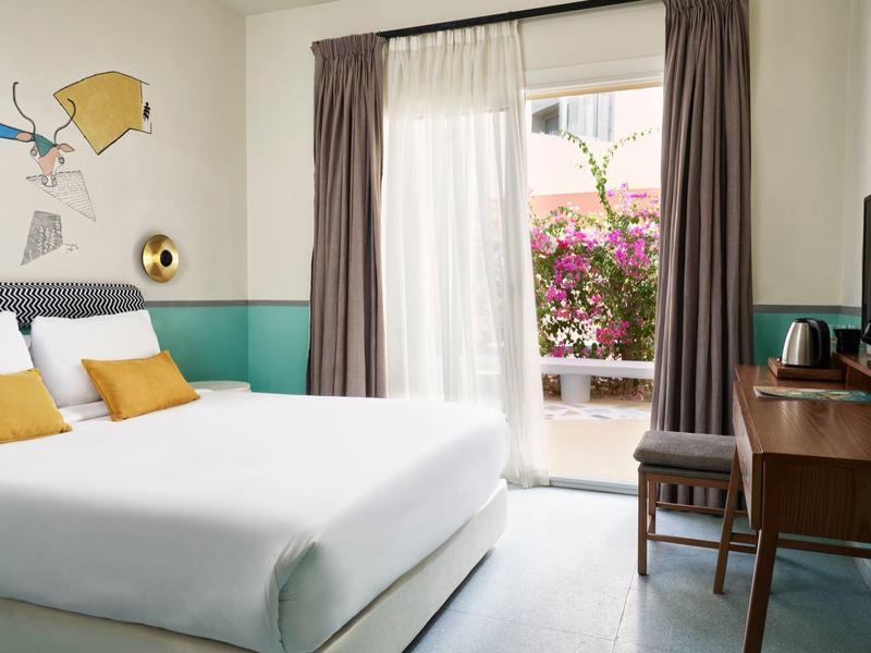 Helles Schlafzimmer mit großem Bett, buntem Wandbild, Schreibtisch, TV und Terrasse mit Blumen.