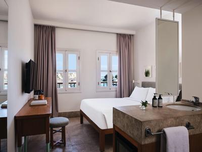 Bright hotel room with double bed, desk, TV, and two windows with curtains.
