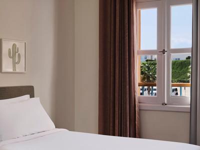 Bright hotel room with white bed, brown curtains, and a view of a balcony with plants.