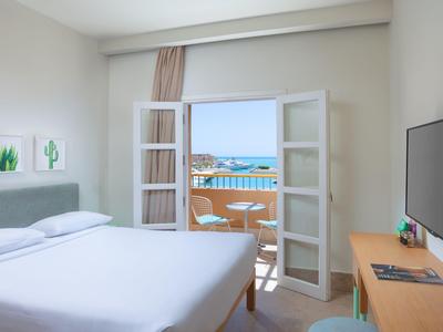 Bright hotel room with bed, desk, and balcony overlooking the sea.