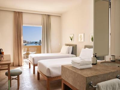 Helles Hotelzimmer mit zwei Einzelbetten, Schreibtisch, Balkon und einem offenen Waschbereich.