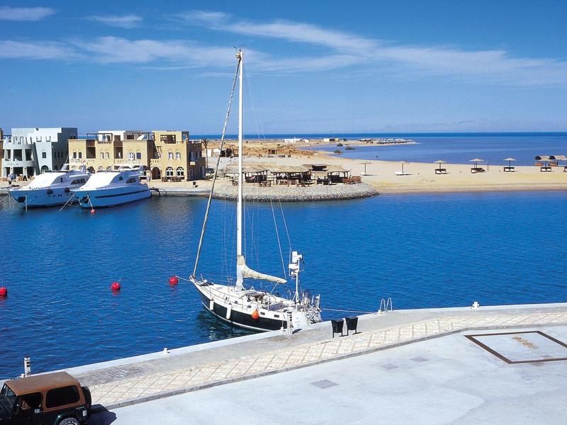 Hafen mit Segelbooten, blauer Wasseroberfläche und gelbem Sandstrand unter blauem Himmel.