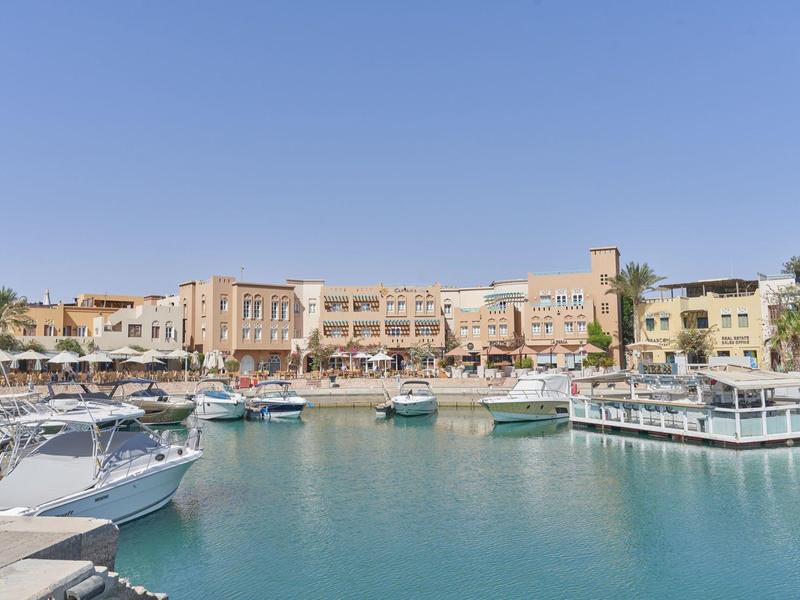 Hafen mit mehreren Booten, türkisem Wasser und sandfarbenen Gebäuden unter klarem Himmel.