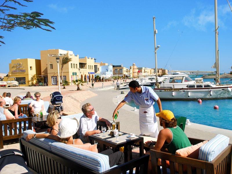 Menschen sitzen in Außenbereich am Wasser, Segelboote und Sandstrand im Hintergrund, sonniger Tag.