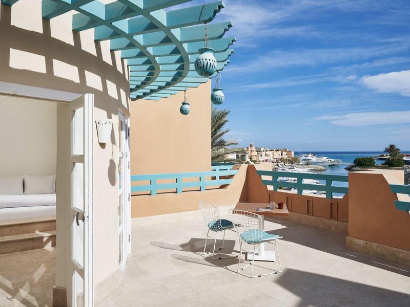 Terrasse mit Stühlen und kleinem Tisch, blauer Himmel, Meer und Gebäude im Hintergrund.